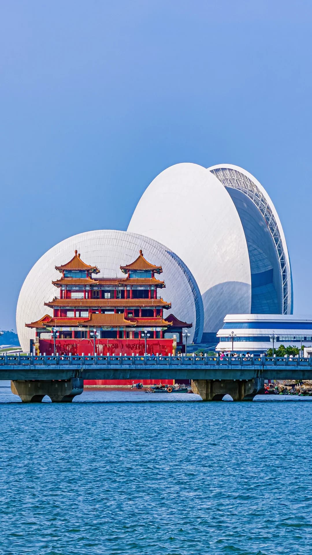 The twin shells of Zhuhai Opera House beyond a red waterfront pavilion and blue sea